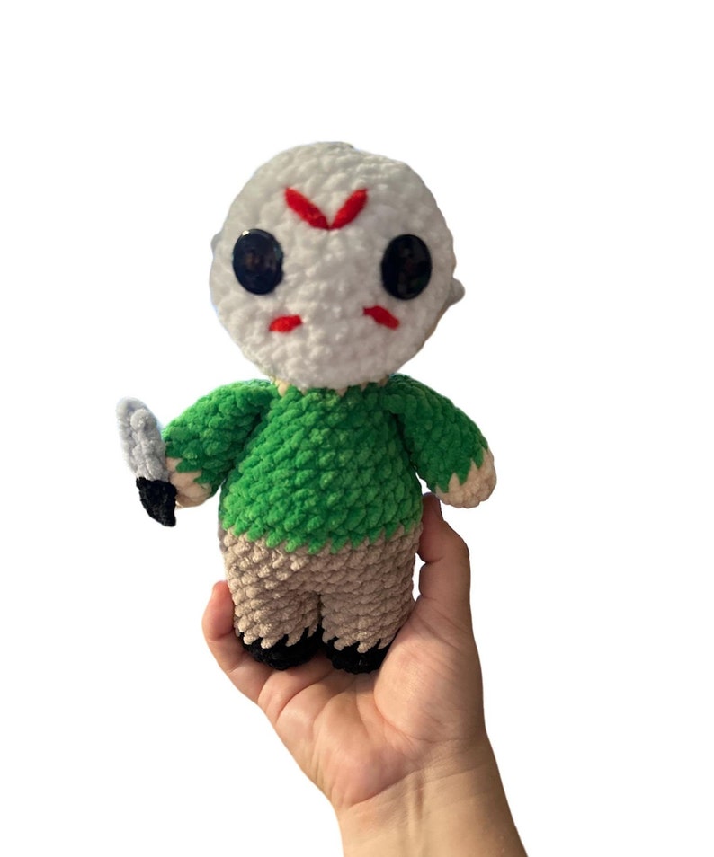 May include: A crocheted plush toy of a horror movie character. The toy is wearing a white mask with red markings and a green shirt. The toy is holding a knife in its right hand.