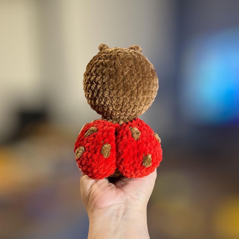 May include: A brown crocheted ladybug toy with red wings and black spots. The toy is being held in a person's hand.