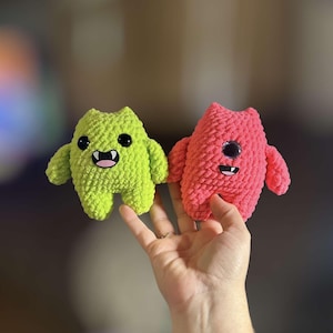 May include: Two crocheted plush toys, one green and one pink, with smiling faces and googly eyes. The toys are held in a hand.