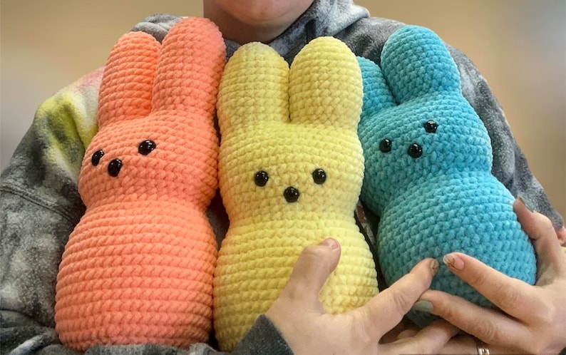 以下が含まれることがあります： ピンク、イエロー、ブルーの3色の編みぐるみウサギのぬいぐるみ。おもちゃはすべて、長い耳と丸い体を持つウサギの形をしています。