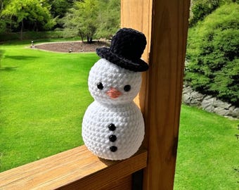 May include: A white crocheted snowman with a black top hat and black button eyes. The snowman is sitting on a wooden railing with a green grassy background.