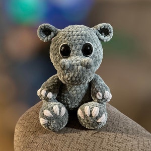 Puede incluir: Un hipopótamo de peluche gris tejido a crochet con grandes ojos negros y detalles blancos en sus pies. El hipopótamo está sentado sobre una superficie gris texturizada.