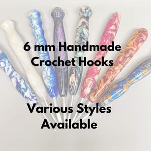 May include: A collection of 6 mm handmade crochet hooks in various styles. The hooks feature colorful, marbled handles in shades of blue, white, pink, purple, and gold. Text on the image reads "6 mm Handmade Crochet Hooks, Various Styles Available."