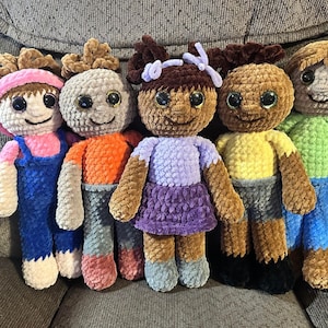 May include: Six crocheted plush dolls with different colored clothing and hair. The dolls are sitting on a gray couch.