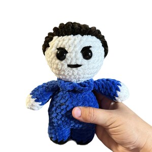 May include: A crocheted plush toy with a white face, black hair, and a blue jumpsuit. The toy has a grumpy expression with black eyes and a frown.