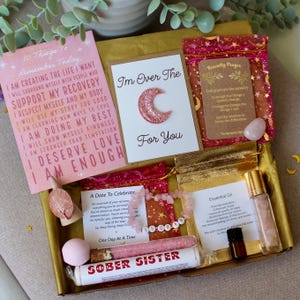 May include: A self-care gift box containing cards with motivational quotes, a bracelet, essential oil, a bath bomb, and a tube labelled "SOBER SISTER". The box has a gold interior and is decorated with pink and gold accents. Items are for well-being.