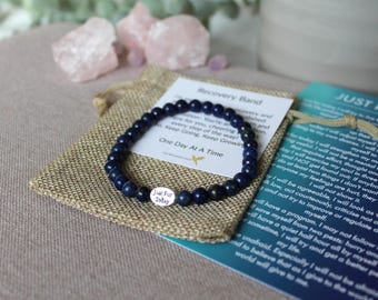 Puede incluir: Una pulsera de cuentas azul oscuro con un pequeño dije plateado que dice "Just For Today" descansa sobre una pequeña bolsa de arpillera. La pulsera está junto a una tarjeta que dice "Recovery Band".