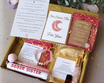 Puede incluir: Una caja dorada llena de artículos de autocuidado, que incluyen tarjetas con afirmaciones, una pulsera, aceites esenciales y una bomba de baño. La caja contiene el texto "I'm Over The C For You" y "Sober Sister". Los artículos son en tonos rosa, dorado y blanco.