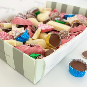 Pode incluir: Uma caixa cheia de uma variedade de doces de chocolate coloridos, incluindo chocolate branco, chocolate ao leite e chocolate preto. Os doces têm uma variedade de formas, incluindo corações, estrelas e redemoinhos.
