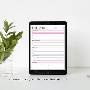 Op de afbeelding: Een zwarte tablet toont een "Brain Dump" sjabloon met roze en blauwe secties voor doelen, thuis, werk, familie, zelfzorg en winkelen. De tekst "customize for your life, download & print" staat onderaan.