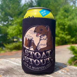 May include: A black foam beverage insulator sleeve featuring a graphic of a brown stoat holding a beer mug. The text reads STOAT STOUT IT'S A MUST(ELID)!. The sleeve is wrapped around a yellow aluminium can with a blue label that says PAS TROP SUCRE.