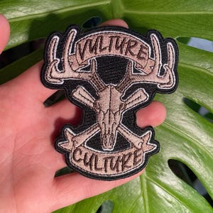 May include: Embroidered patch with a black background and tan accents. The patch features a deer skull with antlers, crossed bones, and the words "VULTURE" and "CULTURE" in a banner design. The patch is held in a hand.