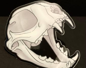 May include: A white and grey detailed illustration of a cat skull with an open jaw and sharp teeth. The skull is outlined in black and set against a black background.