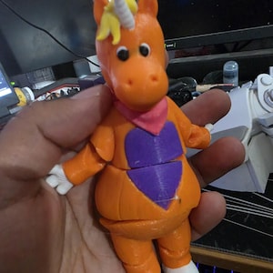 May include: An orange 3D printed unicorn figurine with a yellow mane, a white horn, and white hands and feet. The figure features a pink neck scarf and a purple heart-shaped patch on its chest, with black circular eyes and a smooth, segmented body.