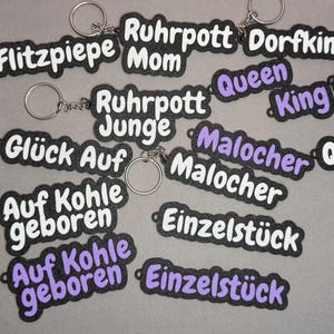 Könnte beinhalten: Eine Sammlung von Schlüsselanhängern mit verschiedenen Textdesigns. Die Schlüsselanhänger haben einen schwarzen Hintergrund mit weißem oder lila Text. Einige Schlüsselanhänger sagen "Ruhrpott", "Queen", "King", "Glück Auf" und "Einzelstück".