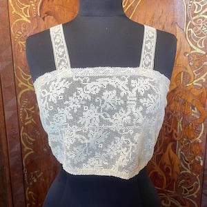 Könnte beinhalten: Ein cremefarbenes Spitzen-Camisole mit quadratischem Ausschnitt und breiten Trägern. Das Kleidungsstück besteht aus einem zarten, durchbrochenen Blumen-Spitzenmuster mit einer Bogenkante am oberen Rand und einem geraden Saum am unteren Ende.