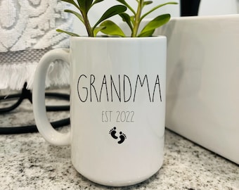 May include: White ceramic mug with black text that reads "Grandma Est 2022" and a pair of black baby footprints. A small green plant is growing in the mug.