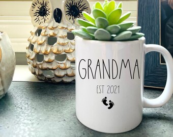 May include: White ceramic mug with black text that reads "Grandma Est 2021" with a pair of black footprints below the text. A succulent plant is in the mug.