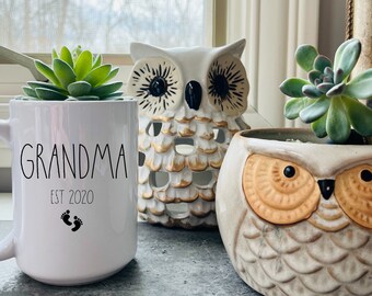 May include: White ceramic mug with the text "Grandma Est 2020" and two black footprints. The mug is sitting on a table next to two owl-shaped planters with succulents.