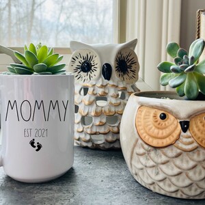 May include: White ceramic mug with the text "Mommy Est 2021" and two black footprints. The mug is sitting on a table next to two owl-shaped planters with succulents.