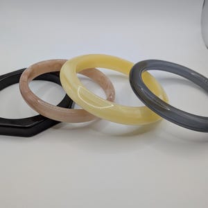 May include: Four colorful bangles in a row. The first is a black, hexagonal shape. The second is a light brown color. The third is a light yellow color. The fourth is a dark gray color.