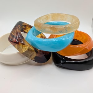 May include: A collection of colorful resin bangles in various shapes. The bracelets include a marbled brown, translucent orange, light blue, black, and a cream-colored bangle. The bangles are stacked on top of each other.