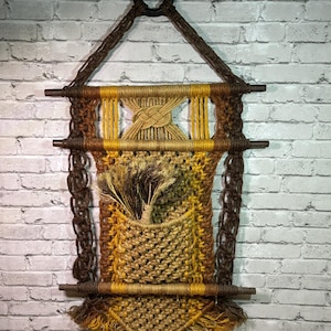 May include: A macrame wall hanging featuring a brown and yellow cord design with three horizontal wooden dowels, a central pocket, and a top black ring. The piece includes intricate knotted patterns, a braided section, and a fringed bottom edge.