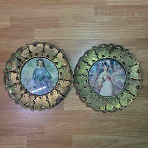 May include: Two antique, round picture frames with ornate, gold-colored leaf and butterfly designs. Each frame holds a vintage portrait of a person. The frames are displayed on a wooden surface.