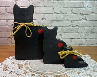 May include: Three black wooden cat figurines of varying sizes, each with a red heart and tied with a natural twine bow. The cats are arranged on a white lace doily, set against a brick wall background.
