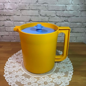 May include: A yellow plastic pitcher with a blue lid and handle. The pitcher has a cylindrical shape and a spout for pouring. The lid has a small, blue knob. The pitcher is sitting on a white lace doily.