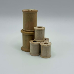 May include: A collection of unfinished wooden spools in various sizes. The spools are cylindrical with a hole through the center, and are stacked and arranged on a white background. The wood has a natural, light brown color.