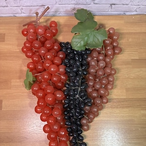 May include: Artificial grape clusters in red, black, and light purple hues. The grapes are arranged with green leaves and brown stems, set against a wooden surface. A decorative item for home decor or crafting.