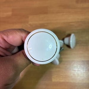 May include: A round white cabinet knob with a smooth, glossy finish and a thin gold-colored circular accent line near the edge. The knob features a central dome shape and includes a metal screw attachment extending from the back for installation.