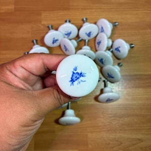 May include: A collection of round white ceramic cabinet knobs, each featuring a small blue floral pattern painted on the front surface. The knobs have a smooth, glossy finish and a metal screw post extending from the back for mounting purposes.