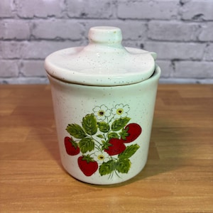 May include: A cream-colored ceramic canister with a matching lid, featuring a speckled finish and a decorative illustration of red strawberries, green leaves, and small white flowers centered on the front of the cylindrical container.