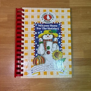 May include: A spiral-bound cookbook titled "Welcome Home For The Holidays" with a snowman illustration. The cover features a yellow plaid pattern and the Gooseberry Patch logo. The book includes holiday recipes, tips, and traditions.