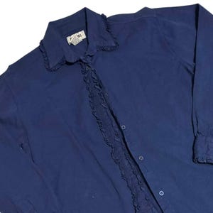 May include: A navy blue button-down shirt with a ruffled collar and front detail. The long-sleeved shirt has a small tag near the collar. The shirt has buttons down the front and on the cuffs.