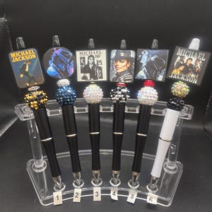 May include: Six pens featuring Michael Jackson themed toppers and decorative rhinestone beads in various colors. The pens have black or white barrels and are displayed in a clear plastic stand labeled with numbers one through six at the base.