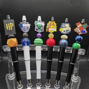 May include: A collection of six decorative pens featuring various designs. The pens have black or white barrels, silver-colored tips, and colorful beaded accents. Each pen is topped with a charm representing a different house from the Harry Potter series, including the HP logo.
