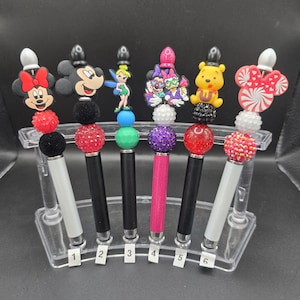 May include: A collection of six decorative pens featuring Disney character toppers. Each pen has a unique design, including Minnie Mouse, Mickey Mouse, Tinkerbell, and Winnie the Pooh. The pens have black or white barrels and are displayed in a clear plastic stand.
