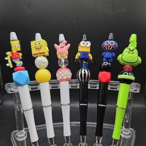 May include: A set of six decorative pens with character toppers, including Bart Simpson, SpongeBob, Patrick Star, a Minion, a blue monster, and the Grinch. The pens have white, black, and green barrels, and are displayed in a clear plastic stand.