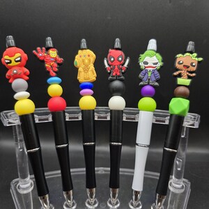 May include: Six pens with superhero-themed toppers, including Iron Man, Thanos, Deadpool, and the Joker. The pens have black or white barrels with silver accents and colorful beads. They are displayed in a clear plastic stand.