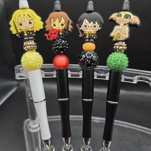May include: Four decorative pens featuring characters from a popular fantasy series. Each pen has a black barrel, a character-themed topper, and decorative beads. One pen has a white barrel. The toppers include a blonde girl, a girl with brown hair, a boy with glasses, and a green-skinned creature.