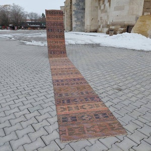 May include: A long, narrow, handwoven rug with a colourful geometric pattern in shades of brown, orange, purple, and blue. The rug is laid out on a grey brick surface.