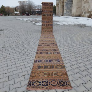 Könnte beinhalten: Ein langer, rechteckiger Läuferteppich mit einem geometrischen Muster in Braun-, Blau- und Orangetönen. Der Teppich wird im Freien auf einer gepflasterten Oberfläche präsentiert und zeigt seine Länge und sein Design.