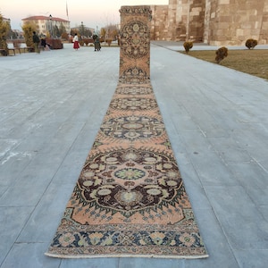 Puede incluir: Una alfombra larga y estrecha con un diseño persa tradicional. La alfombra tiene un fondo rosa claro con acentos marrones oscuros, verdes y crema. La alfombra está colocada sobre un suelo de hormigón gris.