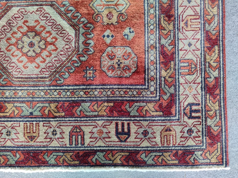May include: Close-up of a red, beige, and blue patterned rug with intricate designs. The rug has a fringe border and a white backing.