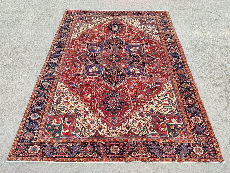 May include: A traditional Persian rug with a red, cream, and blue color scheme. The rug features a central medallion with intricate floral and geometric patterns. The rug has a dark blue border with a repeating pattern.