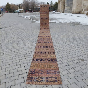May include: A long, narrow, handwoven rug with a geometric pattern in shades of brown, orange, blue, and pink. The rug is laid out on a gray brick walkway.