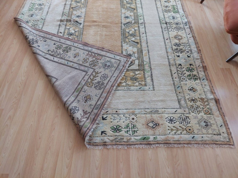 Puede incluir: Una alfombra beige con un patr&oacute;n geom&eacute;trico en tonos de marr&oacute;n, verde y crema. La alfombra tiene un borde de flecos y est&aacute; doblada por la mitad.